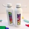 Two white reusable water bottles with colorful vertical lettering and loop caps, surrounded by bright school supplies on a clean desk.
