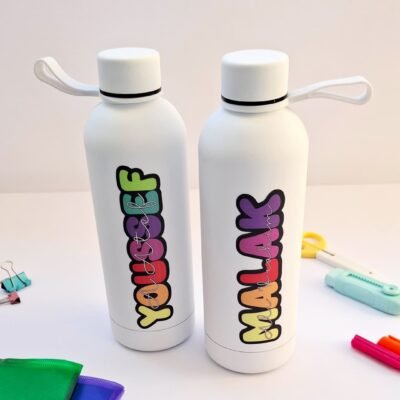 Two white reusable water bottles with colorful vertical lettering and loop caps, surrounded by bright school supplies on a clean desk.