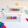 Two clear pencil cases labeled “YOUSSEF” and “MALAK” sit on a white desk with colorful stationery, scissors, clips, and pens, playful tone.