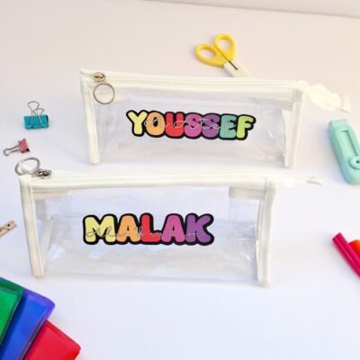 Two clear pencil cases labeled “YOUSSEF” and “MALAK” sit on a white desk with colorful stationery, scissors, clips, and pens, playful tone.