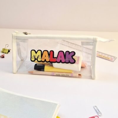Transparent pencil case labeled “MALAK” in colorful letters, holding pens and pencils on a white desk beside notebooks and paper clips.
