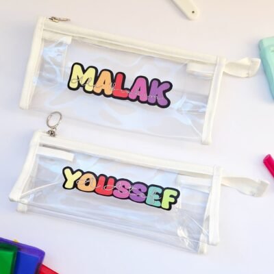Two clear zippered pencil pouches labeled “MALAK” and “YOUSSEF” lie on a white surface, surrounded by colorful stationery, creating a playful mood.