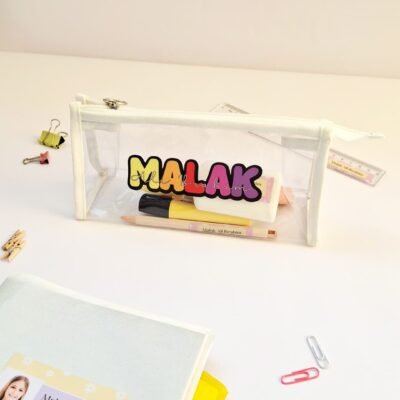 Bright white desk with a transparent pencil pouch labeled “MALAK,” colorful pens, clips, and a decorated notebook; neat, cheerful study scene.