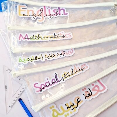 Stack of transparent zippered school files labeled Science, English, Mathematics, Islamic Studies, Social Studies, and Arabic, with pens.