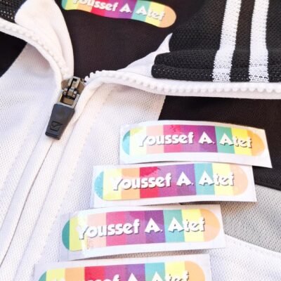 Close-up of a black-and-white jacket with five colorful name labels reading “Youssef A. Atef,” conveying a playful, personalized tone.