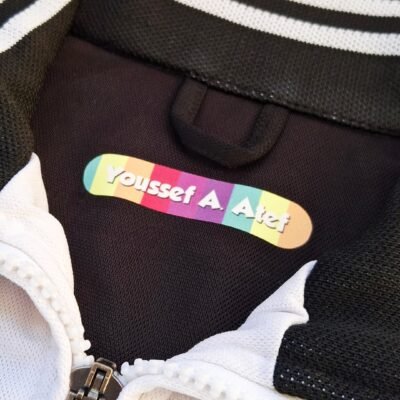 Close-up of a black-and-white jacket with a colorful name tag reading “Youssef A. Atef,” zipper, and textured fabric; playful tone.