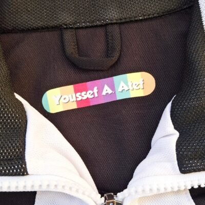 Close-up of a black-and-white zip-up jacket with a colorful “Youssef A. Atef” name patch, textured fabric, and silver zipper.