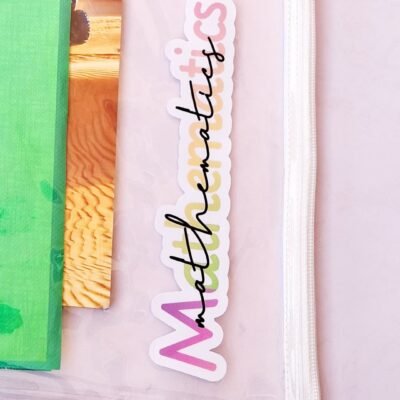 Close-up of a pastel a transparent file with a colorful “Mathematics” sticker, green copybook, and a warm wood-patterned book inside it.