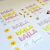 Colorful handmade name labels with cursive names, bright yellow and pink block letters, and smiling sun stickers lie on a white table with clips.